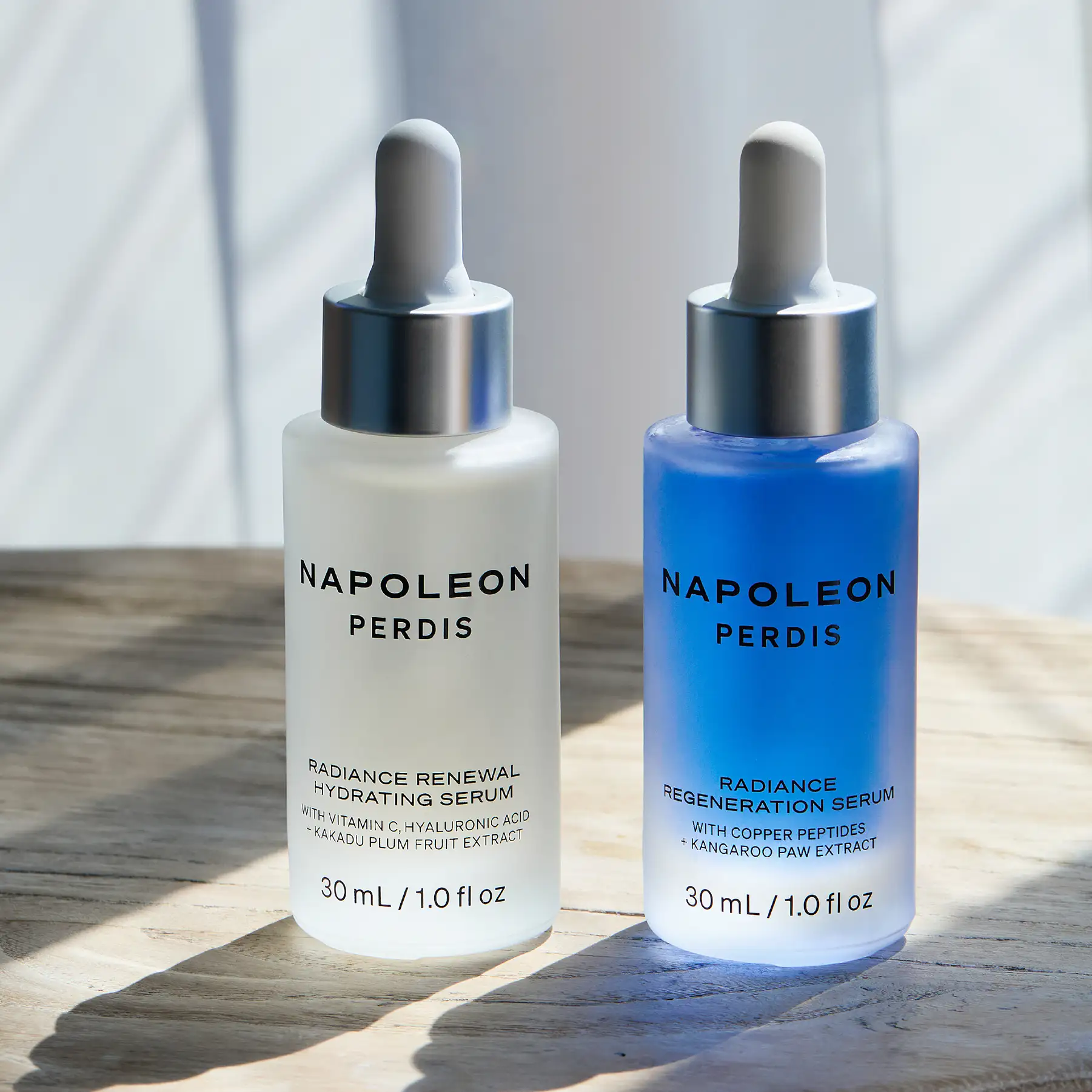 Two Napoleon Perdis serum bottles side by side on a timber surface in dappled light