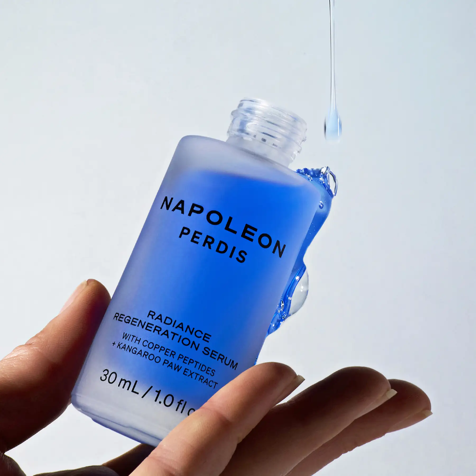 Napoleon Perdis Radiance Regeneration Serum held in the palm of a hand, with a drop falling from the dropper