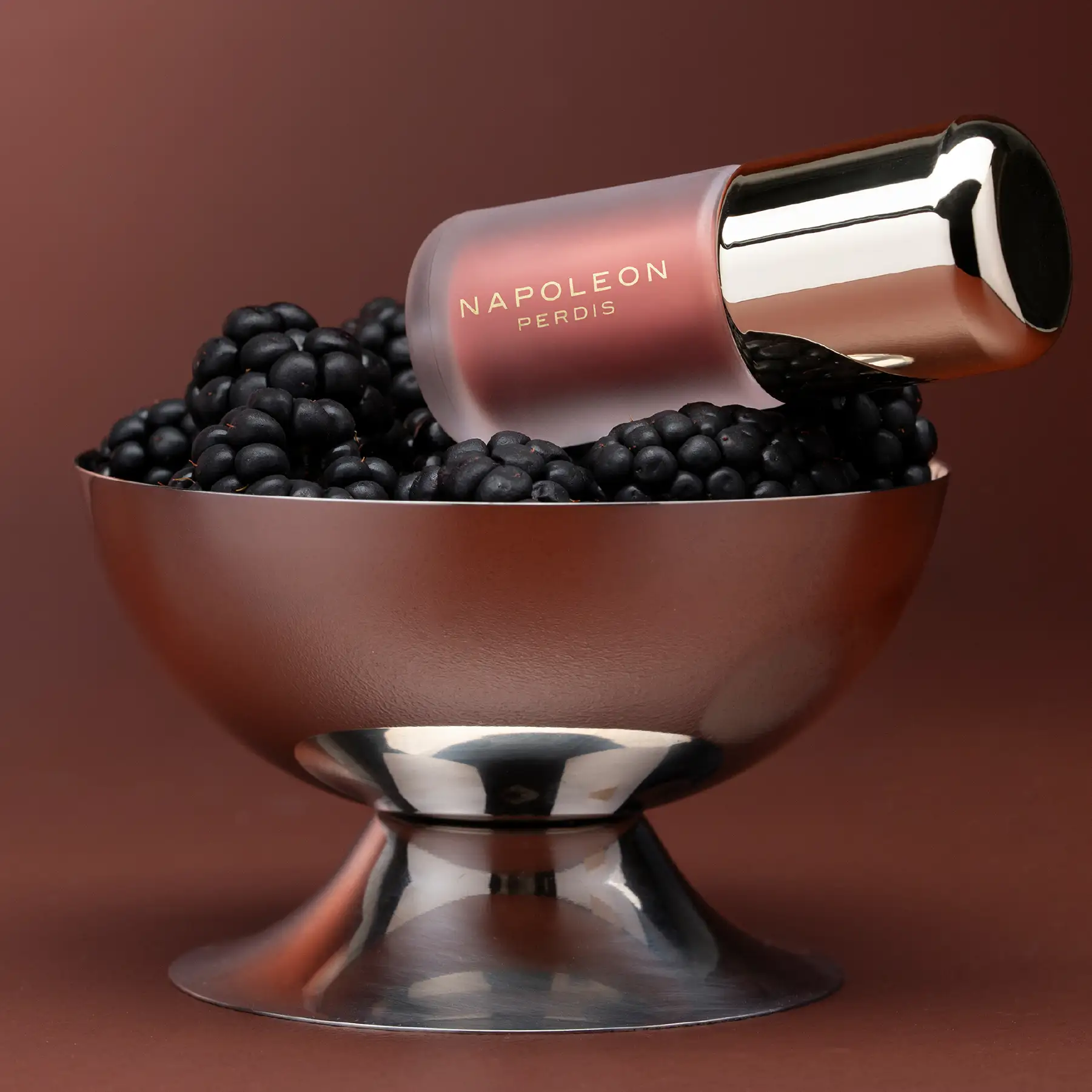 Napoleon Perdis lip gloss balanced on a silver coupe filled with blackberries
