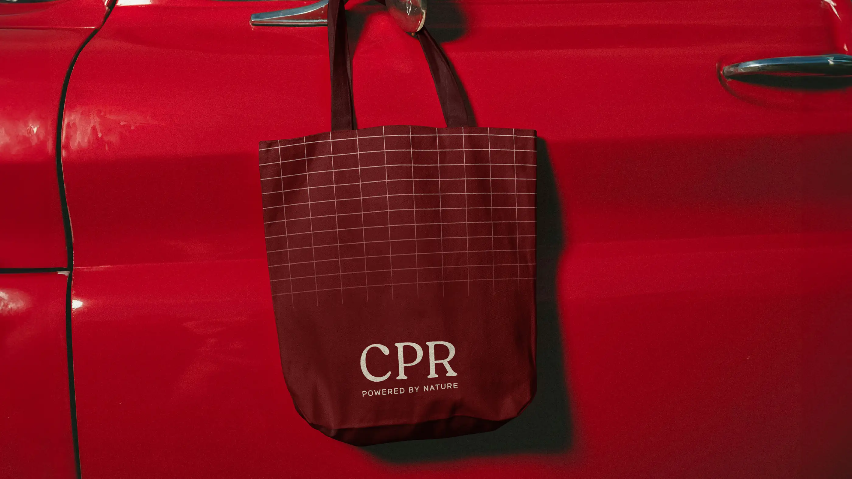 CPR Hair tote bag in deep red with a fine grid pattern, hanging on a red car door